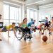 Des lycéens et des lycéennes, dont une sportive en fauteuil roulant, réalisent un circuit de fitness inclusif dans un gymnase lumineux, entre échauffement, gainage et dribbles, dans une ambiance motivante.