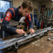 Jeune skieur alpin français en tenue de compétition préparant ses skis dans un local d’équipe de montagne, avec un ordinateur affichant des graphiques budgétaires abstraits.